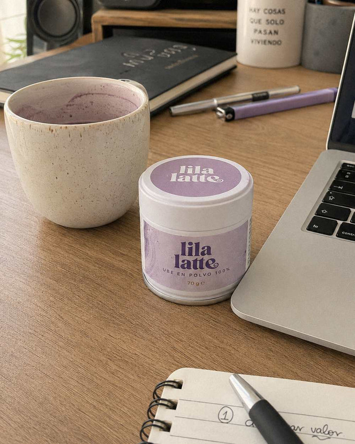 Lata de Lila Latte en un escritorio junto a una taza de latte morado